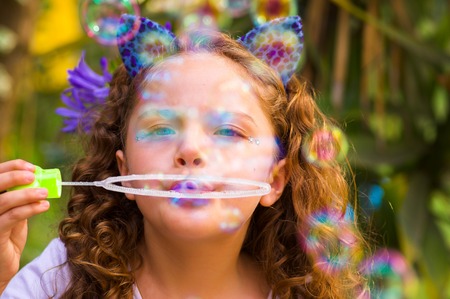 Portrait of a happy little curly girl playing with soap bubbles on a summer nature, wearing a blue ears of tiger accessories over her head in a blurred nature backgroundの写真素材