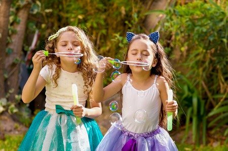 Two happy beautiful little girls playing with soap bubbles on a summer nature, one girl is wearing a blue ears tiger accessories over her head and both girls wearing a princess dress in a blurred nature backgroundの写真素材