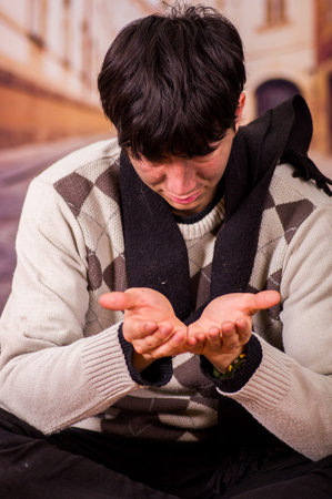Close up of a homeless young man in the streets, asking for money with both hands, in a blurred...