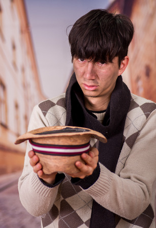 Close up of a sad homeless young man in the streets, with a hat in his hands, asking for money, in a blurred backgroundの写真素材