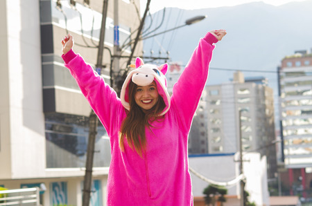 Close up of a beautiful smiling young woman wearing a pink unicorn costume, with both hands up at outdoors in the city of Quitoの写真素材