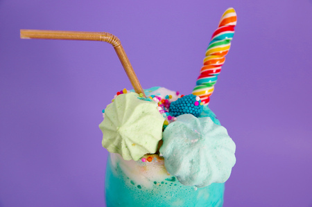 Close up of a delicious homemade extreme milkshake with dragees over a milk foam and a rainbow candy on top with a plastic straw in a purple backgroundの写真素材
