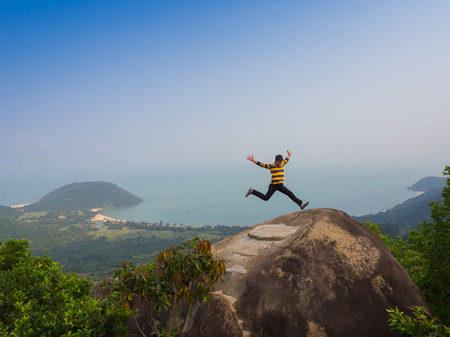 HOIAN, VIETNAM, SEPTEMBER, 04 2017: Unidentified man jumping over a huge rock with a beautiful view of the coastline of Danang, Vietnam, in a sunrise from the top of a mountainのeditorial素材