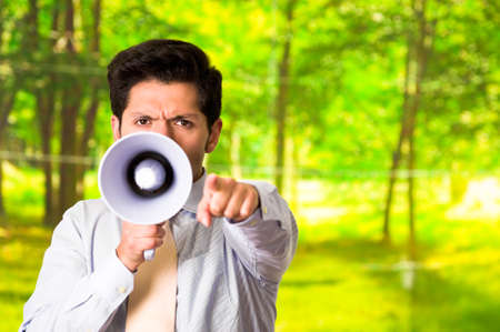 Portrait of a handsome man shouting with a megaphone while he is pointing in front of him his hand, in a blurred green backgroundの写真素材