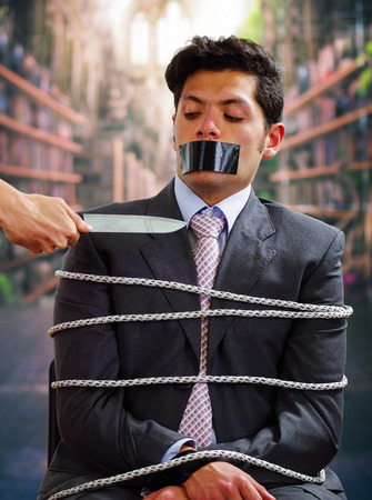 Businessman trapped in a chair with rope, with a black tape in his mouth, afraid of a hand that is holding a knife near of his neack, in a blurred backgroundの写真素材