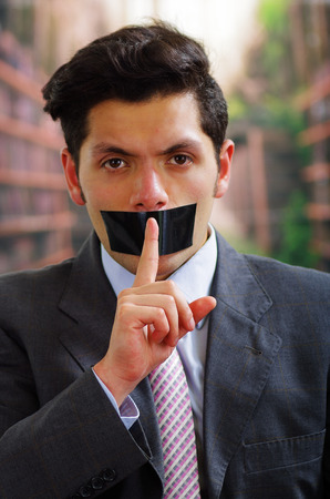 Businessman, with a black tape in his mouth doing a silence sign with a finger in from of his mouth, in a blurred backgroundの写真素材
