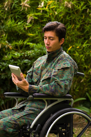 Close up of a handsome soldier sitting on wheel chair using his tablet with both hands, and wearing military uniform in a nature backgroundの写真素材