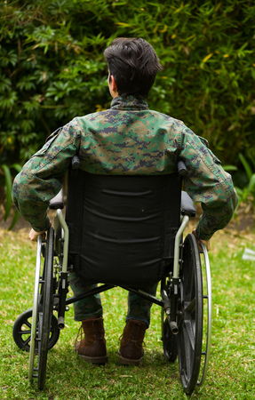 Young soldier sitting on wheel chair at outdoors in the patio, touching with his hands the wheels and giving a back, in a backyard backgroundの写真素材
