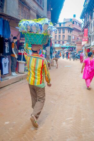 BHAKTAPUR, NEPAL - NOVEMBER 04, 2017: Unidentified man carrying over his head a basket with spoons at outdoor of old building close to markets, with tourists walking around in Bhaktapur, Nepalのeditorial素材