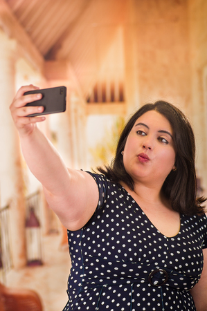 Young beautiful plus size model in black dres, taking herslef a selfie with her cellphone, xxl woman on blurred studio backgroundの写真素材