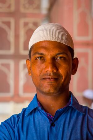 Delhi, India - September 27, 2017: Portrait of a handsome man wearing a blue t-shirt and a white indian hat, looking at camera inside of the temple in Jama Masjid Mosque in Delhi, India in a blurred backgroundのeditorial素材
