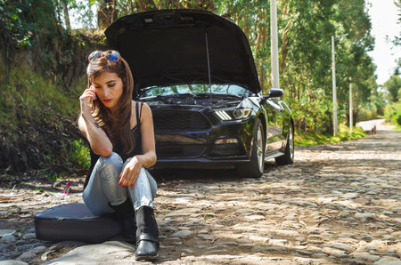 Close up of worried woman sitting in the ground using her cellphone and wearing sunglases in her head, black t-shirt and jeans, having a problem with black car on a roadtrip, the car standing on the sidelines. The hood is openedの写真素材