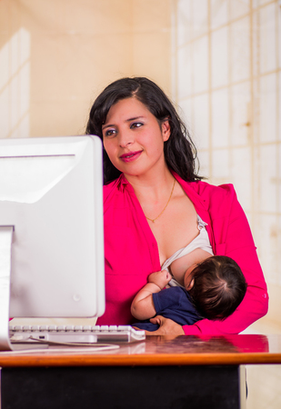 Young beautiful businesswoman working in the office while sitting with his baby boy at her working place, breastfeeding in a blurred backgroundの写真素材
