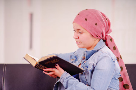 Close up of a woman with cancer wearing headscarf,sitting in a black couch reading a book, in a...