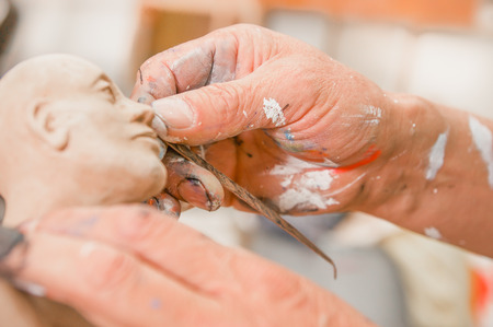 Close up of man ceramist hands holding a tool and working on sculpture details of the head on wooden table in workshopの写真素材