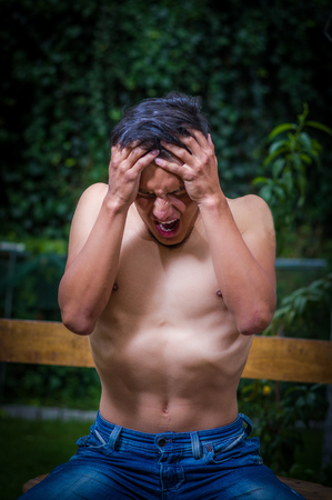 Close up of shirtless man suffering from anorexy, screaming with both arms around her head suffering pain, in a blurred backgroundの写真素材