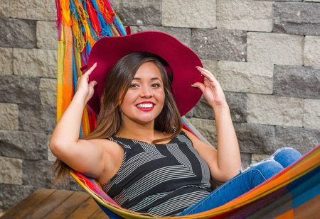Close up of young beautiful woman relaxing in a hammock and wearing a red fashion hat holding with...