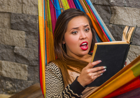 Close up of surprised young beautiful woman reading a book in a hammock, in blurred backgroundの写真素材