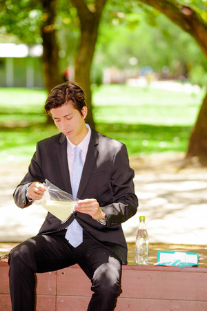 Handsome young businessman wearing a suit and holding a sandwich inside of a plastic bag at outdoors, in a blurred park backgroundの写真素材