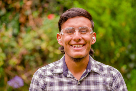 Portrait of smiling young worker wearing transparent safety glasses, long sleeve shirt and ear plugs, in a blurred nature backgroundの写真素材