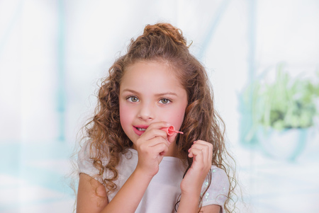 Portrait of beautiful curly little girl painting her nails, wearing a white blouse in a blurred backgroundの写真素材