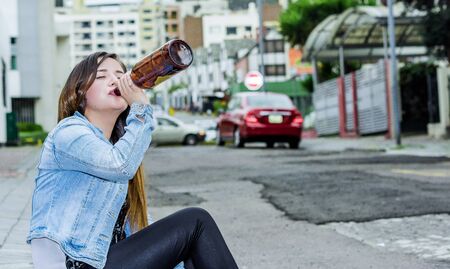 Beautiful young drunk woman sitting in a sidewalk with a bottle of beer in her hand and drinking, desesperate woman and addict conceptの写真素材
