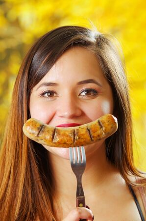Close up of smiling beautiful young woman pointing in front of her mouth a piece of sausage using a fork, looks like a huge smile, BBQ in the garden. Bavarian sausages, in a blurred backgroundの写真素材
