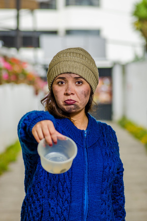 Outdoor view of homeless sad woman on the street in cold autumn weather holding an empty plastic flask in her hands asking for money, at sidewalkの写真素材