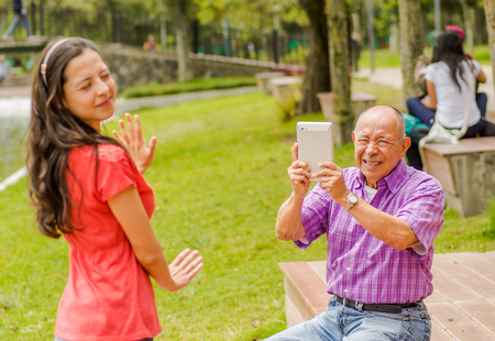 Outdoor view of father taking pictures with a tablet to his daughter while she is trying to avoid the pictures, in the parkの写真素材