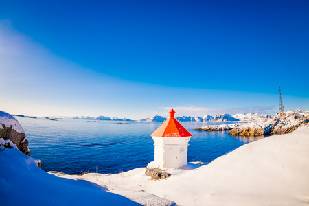 HAMMNOY, LOFOTEN, NORWAY, APRIL, 10, 2018: Outdoor view of white stoned structure with red rooftop in the border and land covered with snow with blue lake in snowy winter in the Arctic Circleのeditorial素材