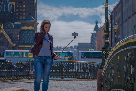 ST. PETERSBURG, RUSSIA, 01 MAY 2018: Blonde russian woman wearing warm clothes taking a selfied using her selfiestick, with blurred background of the city in the horizont in St. Petersburgのeditorial素材