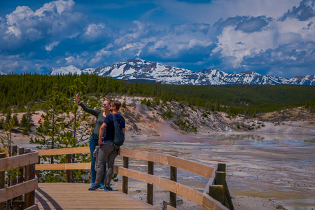 YELLOWSTONE, MONTANA, USA JUNE 02, 2018: Couple taking selfies with pools of colorfully colored water behind in Norris Geyser Basin in Yellowstone with a mountains with snow behindのeditorial素材