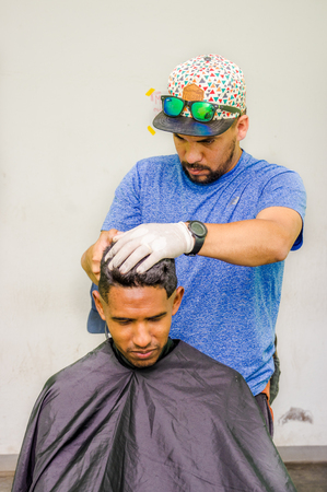 QUITO, ECUADOR, AUGUST 21, 2018: Outdoor view of unidentified man wearing gloves and hat, using a cut hair machine in a youg guy, inside of a refuge for Venezuelan peopleのeditorial素材