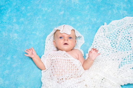 Cute little blue eyed baby looking into the camera lying on the bed wrapped in a knitted blanket