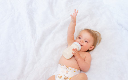 happy little girl drinks milk from a bottle while lying on the bed. Baby food. concept of artificial feed.の写真素材