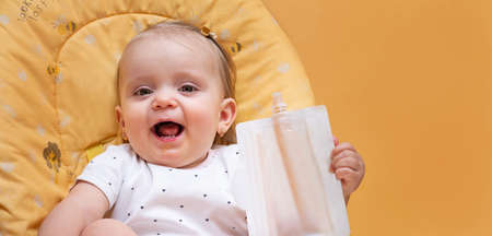 Little toddler girl eats smoothies with a mixture of fruit peach, apple, banana, mango, pumpkin. Sitting in a yellow chair on a yellow background. Baby food.の写真素材