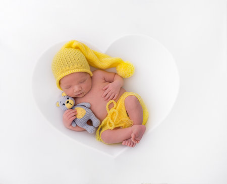 Sleeping newborn baby in the first days of life in yellow pants and a hat with a toy in his hands, white background in the shape of a heart.の写真素材