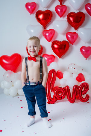 portrait of a blond boy in suspenders and a red bow tie on a white background with heart-shaped balloonsの写真素材