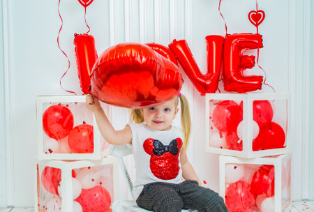 Cheerful little girl holding a red heart balloon on a background of red and white balloons. Valentine's Day.の写真素材