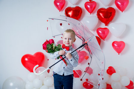 blond boy stands with an umbrella decorated with hearts and a bouquet of roses flowers on Valentine's Day on a white background with red heart-shaped balloons.の写真素材