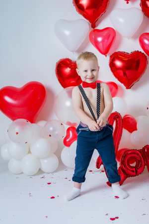 portrait of a blond boy in suspenders and a red bow tie on a white background with heart-shaped balloonsの写真素材