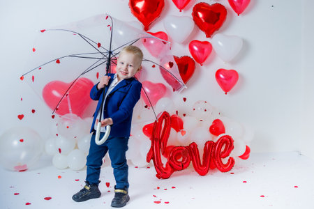 cute boy in classic suit stands with umbrella decorated with hearts on Valentine's Day on white background with red heart-shaped balloons.の写真素材