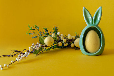 Easter composition on a yellow background. Egg with bunny ears and a branch and Easter eggs with...