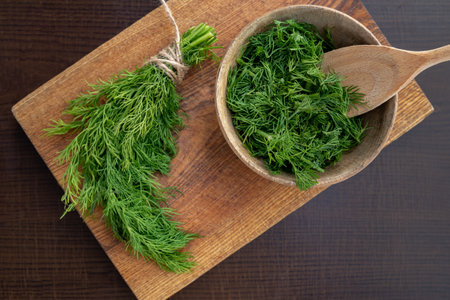 Chopped fresh dill in a wooden plate on a brown kitchen board background next to a bunch of dill. Preparing for freezing. Concept of aromatic herbs for cooking.の写真素材