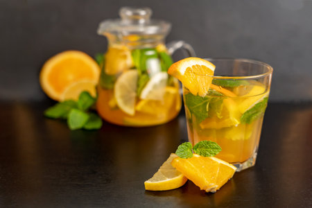 Orange and mint lemonade in a clear glass with orange slices in front on a blurred dark background with a teapot of orange drink behind. Natural refreshment, diet drinks, healthy eating.の写真素材