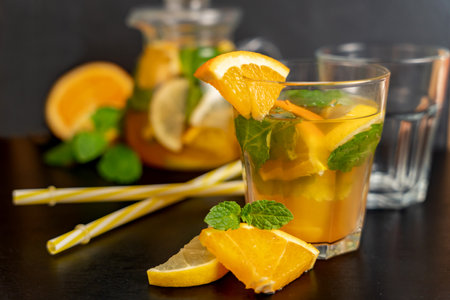 Orange lemonade in a transparent glass with straws and orange slices in front on a blurred dark background with fruits and drink. Summer drinks concept. Natural refreshment, diet drinks, clean eating.の写真素材