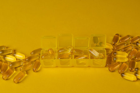 Organizer with yellow capsules of Omega-3 or vitamin D on scattered capsules on a yellow background....