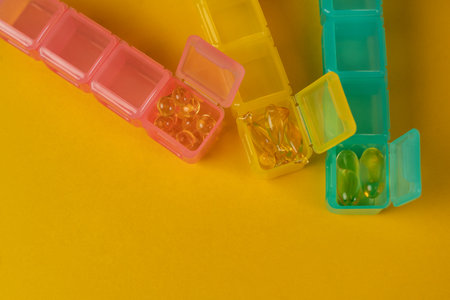 Three colorful organizers with yellow capsules of Omega-3 or vitamin D on a yellow background. Organizers for dietary supplements, pills for every day.の写真素材
