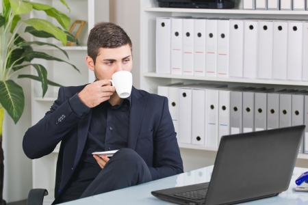  young businessman looking at his laptop holding a coffee sitting at his desk の写真素材