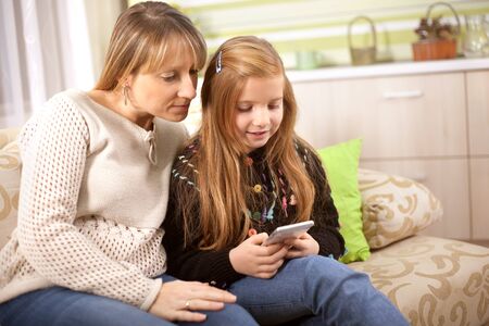 Beautiful young mom and her little daughter are using a smartphone and smiling while sitting on sofa at homeの写真素材